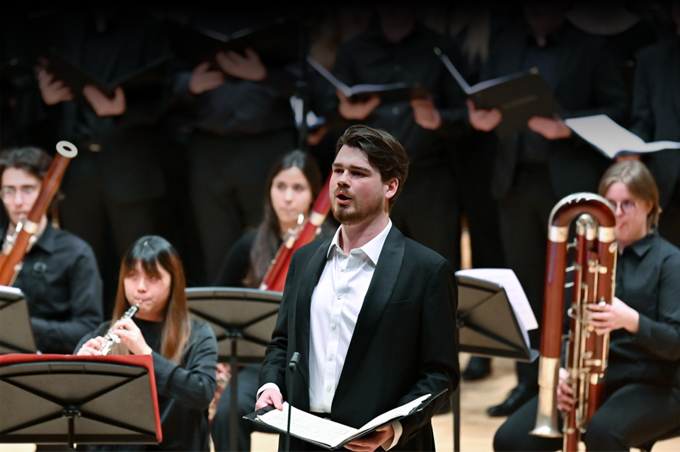 Male student, dressed in a formal attire, performing in an orchestra, with musicians and the choir in the background,